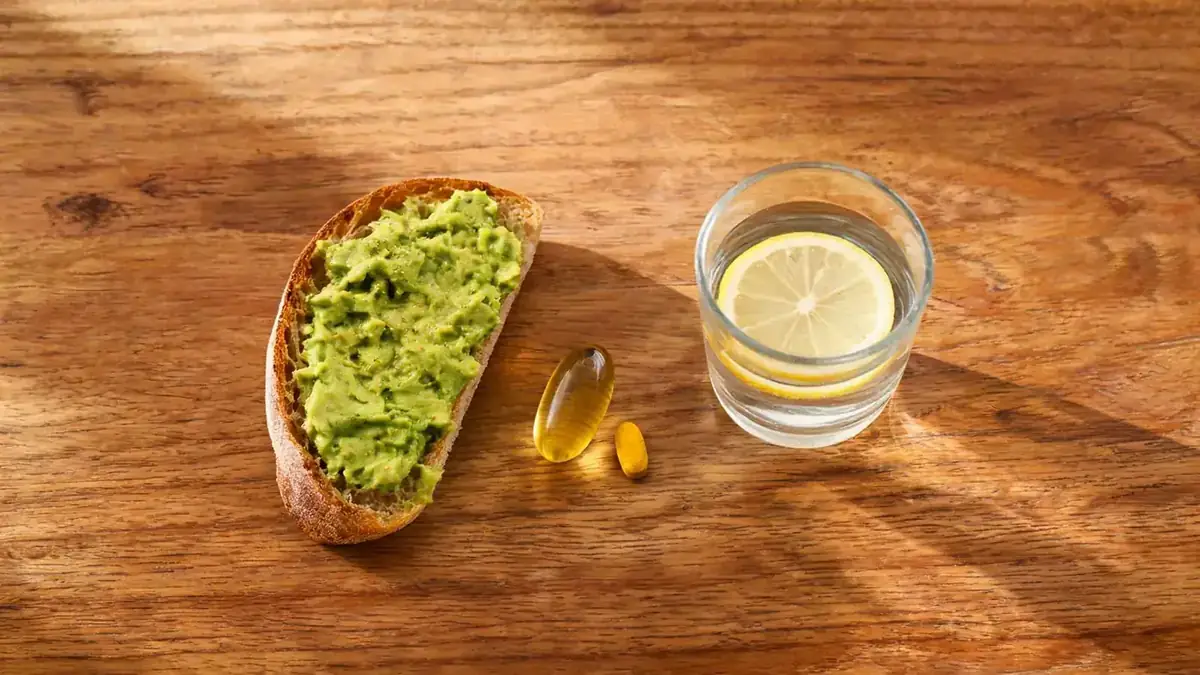 Small breakfast tableau with avocado toast, a fish oil softgel, a turmeric capsule, and a glass of water on a wooden table