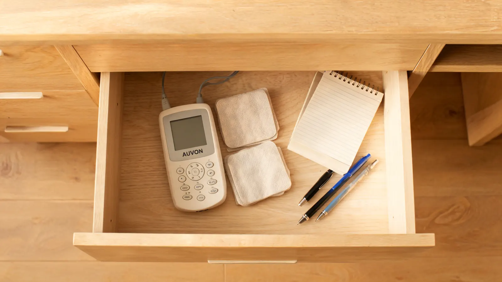 TENS unit and two electrode pads sitting in an open desk drawer lined with a notebook and pens, work-from-home setting