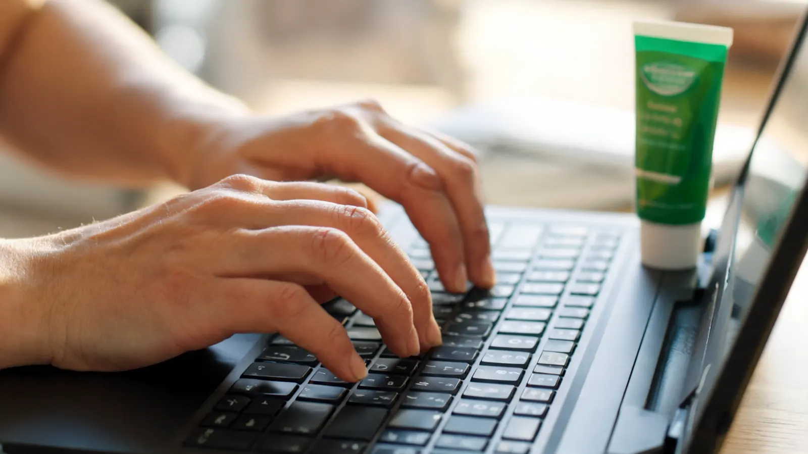 Close-up of two hands with visibly swollen knuckles attempting to type on a laptop keyboard, a green tube of pain cream lying next to the laptop