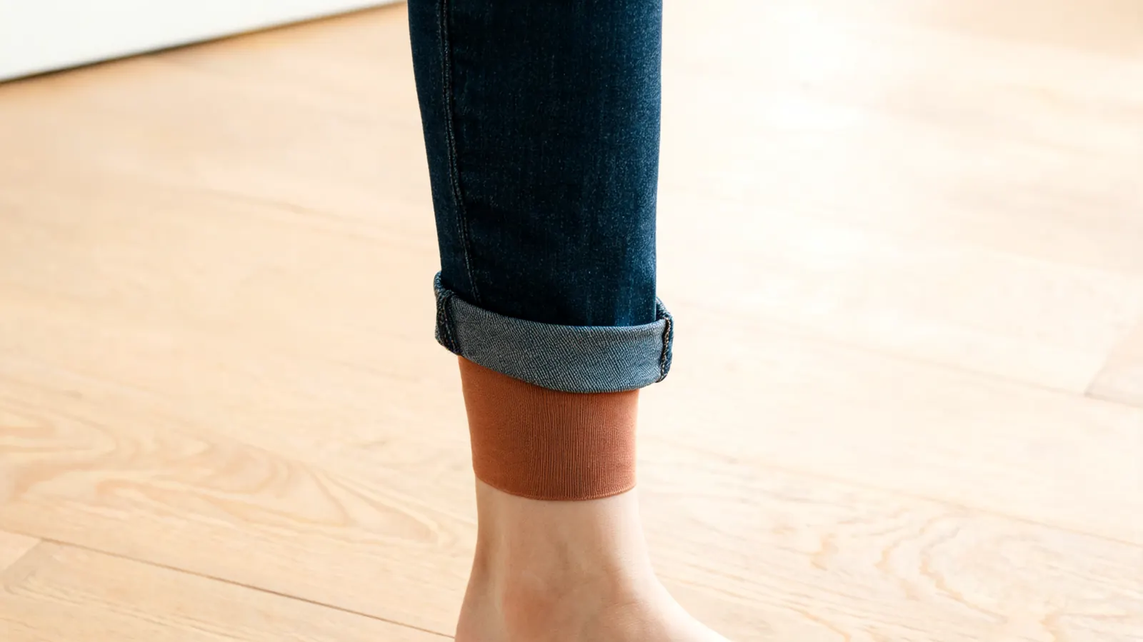 Close-up of a knee compression sleeve barely visible below the cuff of slim dark jeans, standing on a hardwood floor
