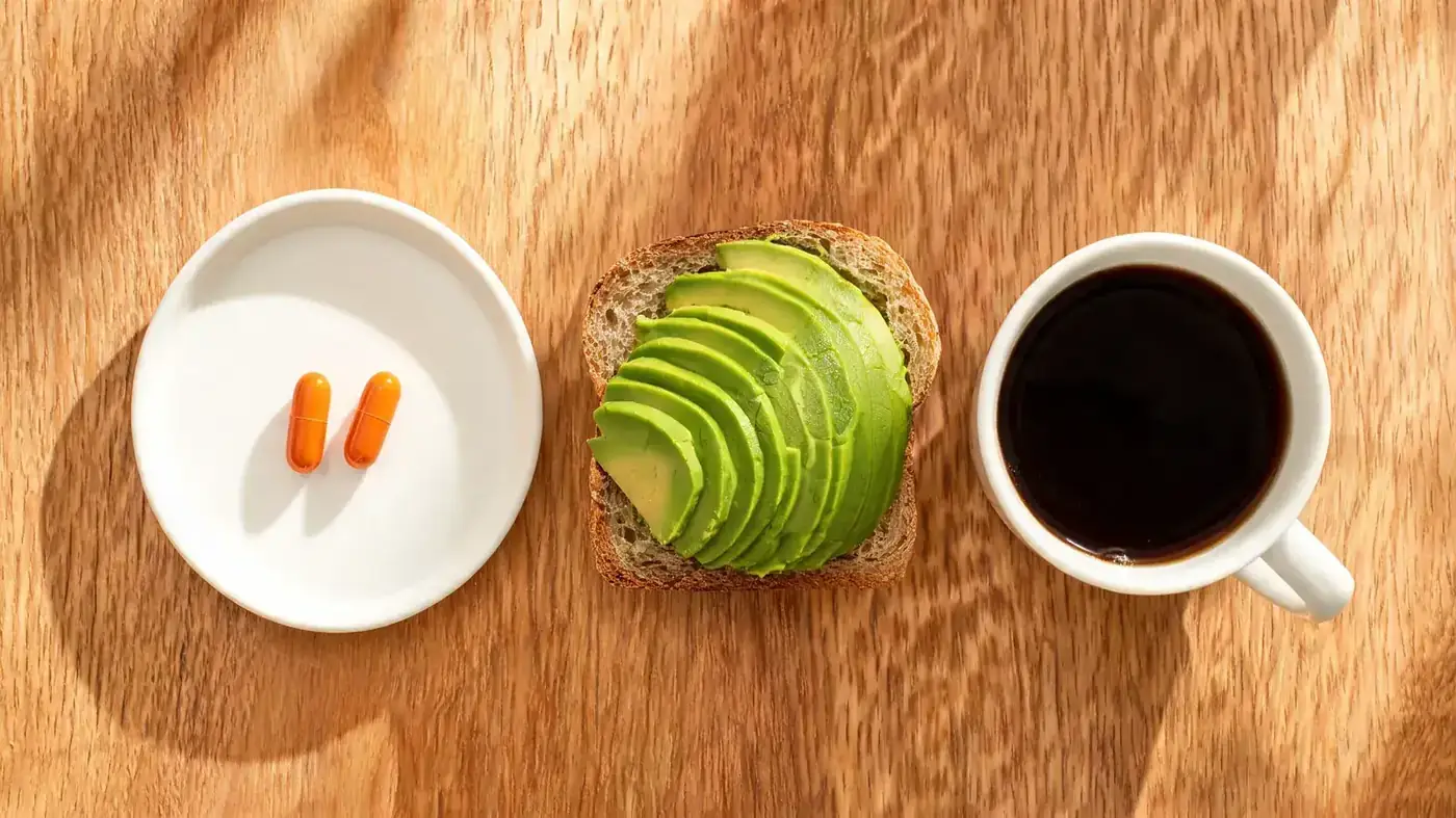 Two orange capsules resting on a small white ceramic plate next to a slice of avocado toast and a ceramic coffee mug on a wooden table
