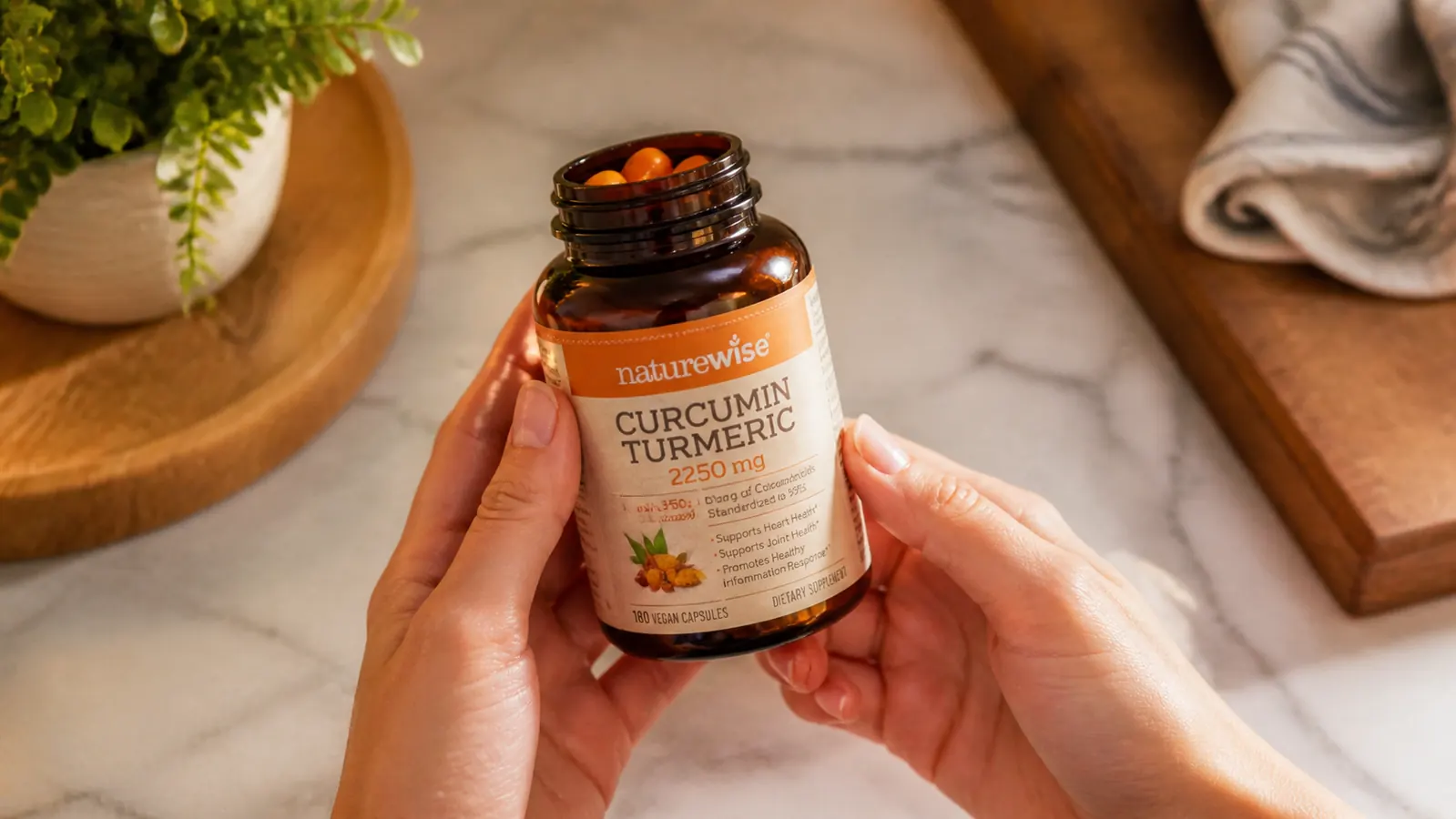 Close-up of hands holding an open bottle of NatureWise Curcumin Turmeric 2250mg capsules, capsules visible inside, natural kitchen light